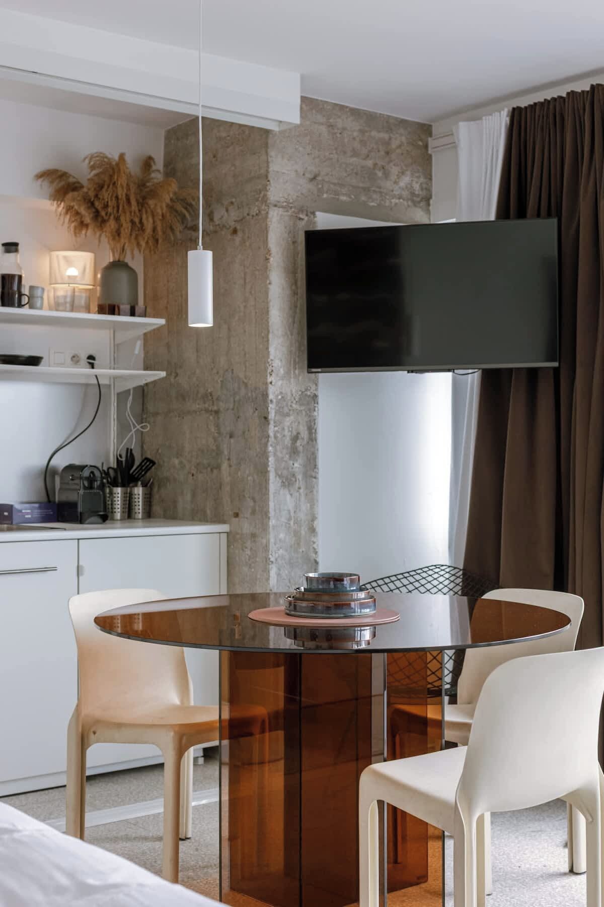 A modern dining area with a glass-top table, white chairs, a flat-screen TV, and a concrete accent wall.