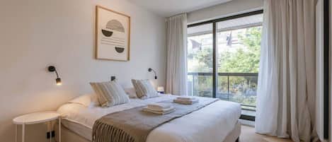 Une chambre à coucher avec un grand lit, une table de chevet, une lampe fixée au mur et une fenêtre avec des rideaux.