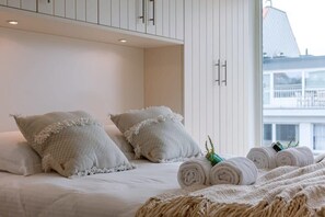 Ein Hotelzimmer mit Bett, weißen Kissen, aufgerollten Handtüchern und Blick auf ein Gebäude durch ein Fenster.