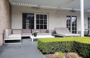 Un espace extérieur aménagé avec un canapé et un fauteuil de relaxation, une table blanche et un arbuste taillé avec soin.