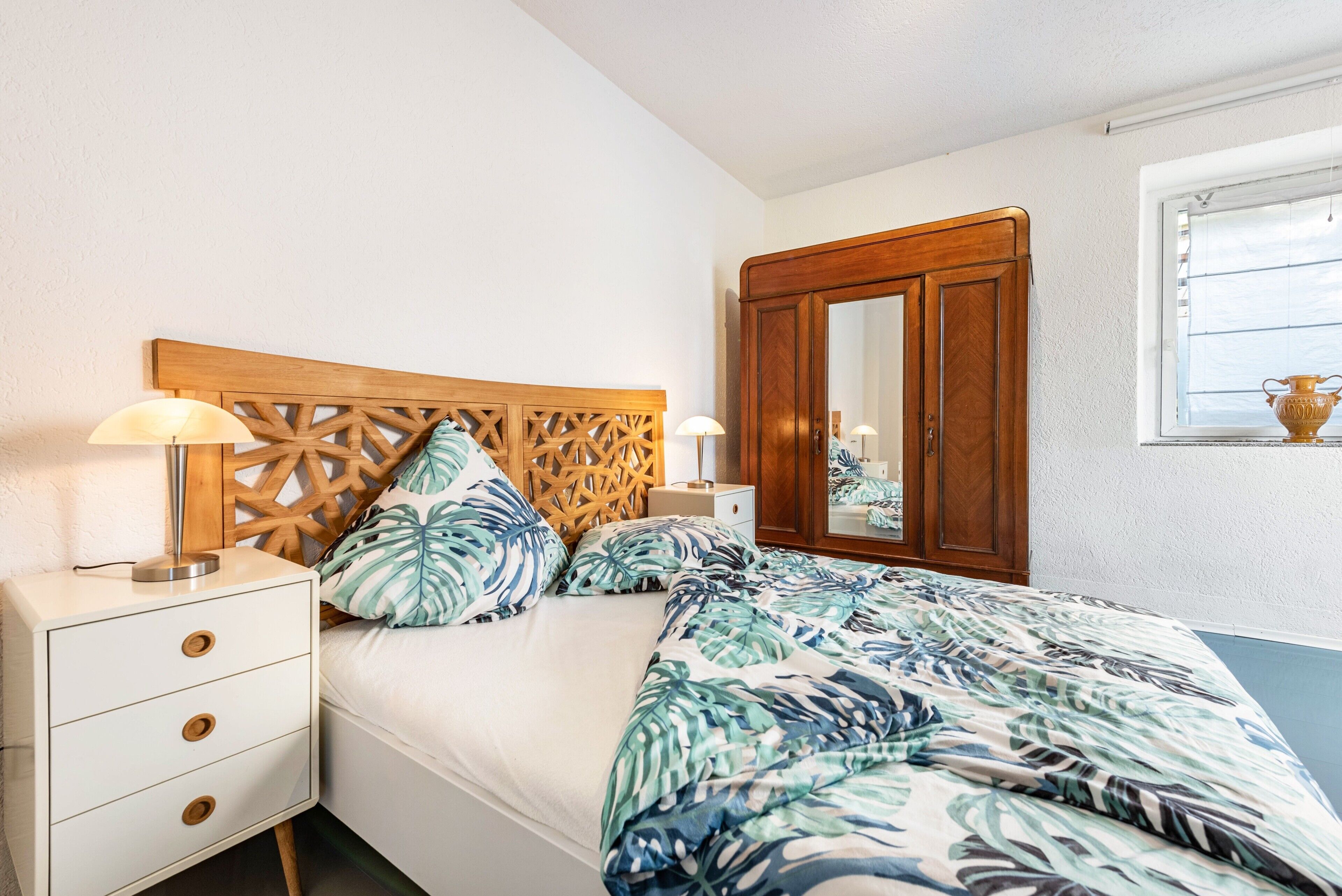 A bedroom with a wooden headboard, a bed with a tropical leaf-patterned bedspread, a nightstand with a lamp, and a wardrobe with a mirror.