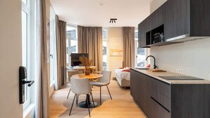 Moderni hotellihuone, jossa on ruokailutila, tummapuukalusteilla varustettu keittiö, taulutelevisio ja suuri ikkuna, jossa on verhot.