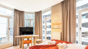 Hotellihuone, jossa on sänky, työpöytä, tuoli, televisio ja suuret ikkunat.