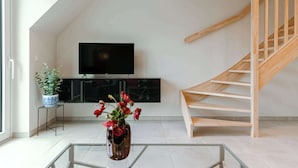 A modern living room with a TV, a potted plant, a vase with flowers, and a wooden staircase. - Beachfront Apt w/Terrace, City View & Fast WiFi (Knokke-Heist)