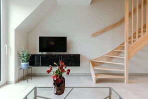 A modern living room with a TV, a potted plant, a vase with flowers, and a wooden staircase. - Beachfront Apt w/Terrace, City View & Fast WiFi (Knokke-Heist)