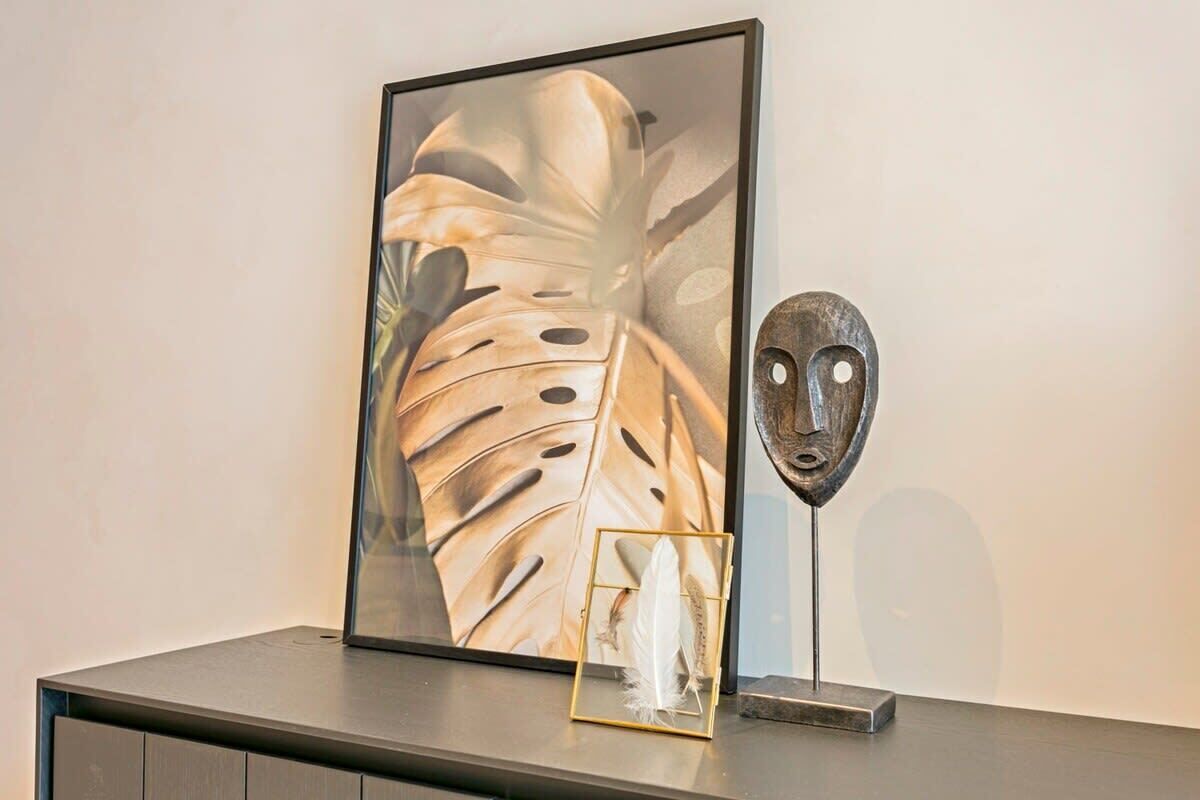 A framed artwork with a tropical leaf pattern, a feather in a glass case, and a sculptural face on a dark surface.