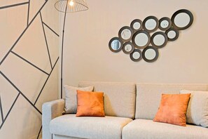 Uma sala de estar moderna com design geométrico na parede, um sofá em tons claros com almofadas laranja e um arranjo circular de espelhos fixados na parede.