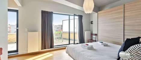 A modern hotel room with a large bed, a ceiling light, a view through a sliding door to an outdoor area, and a radiator on the wall.