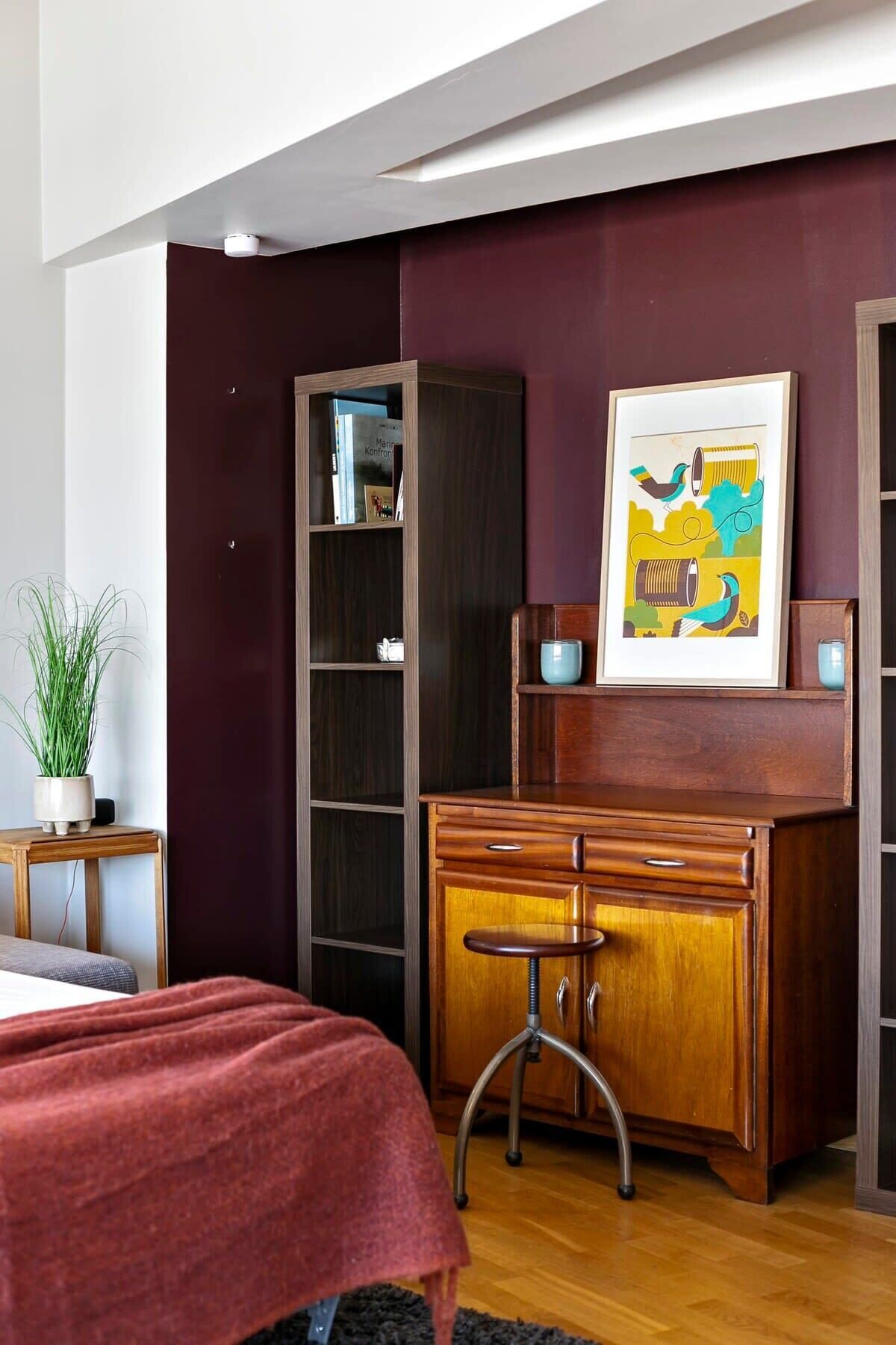 Une chambre à coucher avec un lit, un bureau avec des tiroirs, une bibliothèque, un tableau encadré au mur et une plante en pot sur une table d’appoint.