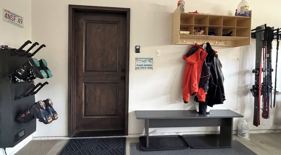 Una stanza con una porta in legno, una panca, un cartello per lo stoccaggio degli scarponi da sci e uno scaffale a parete con attrezzatura da sci.