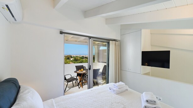 A hotel room with a bed, a TV, a balcony with a table and chairs, and a view of the city. - Azure II Suite (Lefkada)