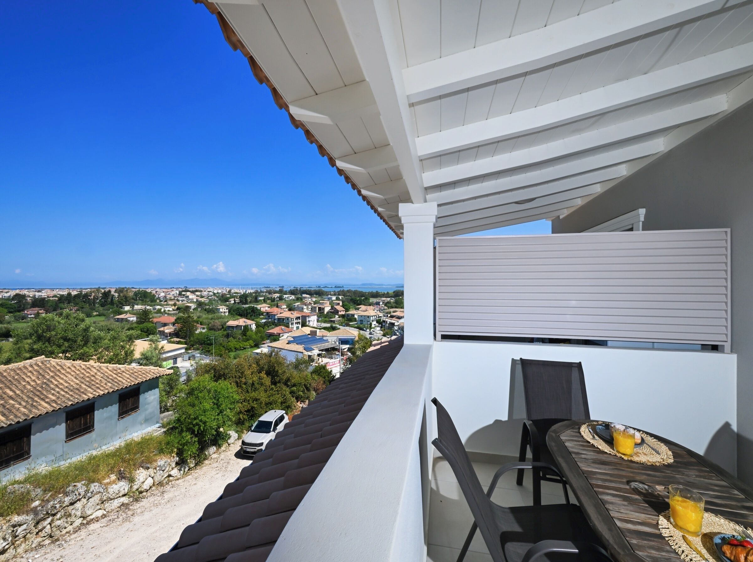 A balcony with a table and chairs, overlooking a residential area with houses and a clear blue sky.