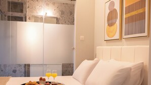 A hotel room with a bed, breakfast tray, artwork, and a shower area. - Azure II Suite (Lefkada)