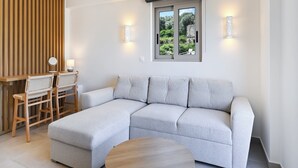 A modern living room with a grey sectional sofa, wooden sideboard, and two chairs. There is a window with a view of greenery and a lamp on the wall. - Azure II Suite (Lefkada)