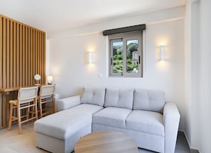 A modern living room with a grey sectional sofa, wooden sideboard, and two chairs. There is a window with a view of greenery and a lamp on the wall. - Azure II Suite (Lefkada)