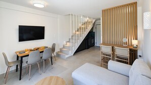 A modern living room with a flat-screen TV, a dining area with chairs and a table, a staircase, and a seating area with a sofa and a small table. - Azure II Suite (Lefkada)