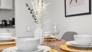 Mesa de jantar posta com tigelas brancas e copos de vinho, um vaso com flores brancas e um jogo americano trançado.