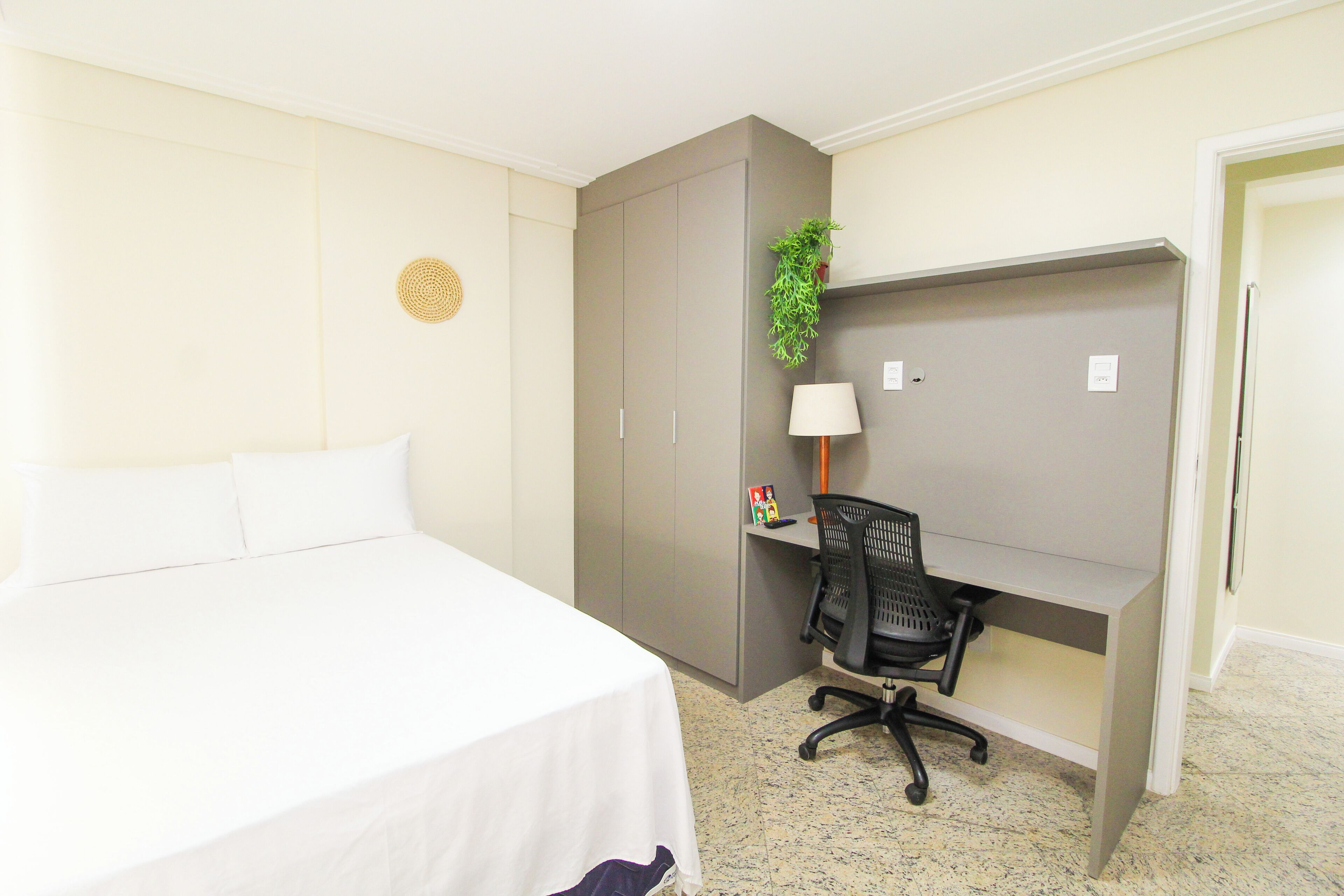 A hotel room with a bed, a desk, a chair, a plant, and a wall decoration.