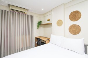 Quarto de hotel com cama, escrivaninha, cadeira, ar condicionado, planta e decoração na parede.