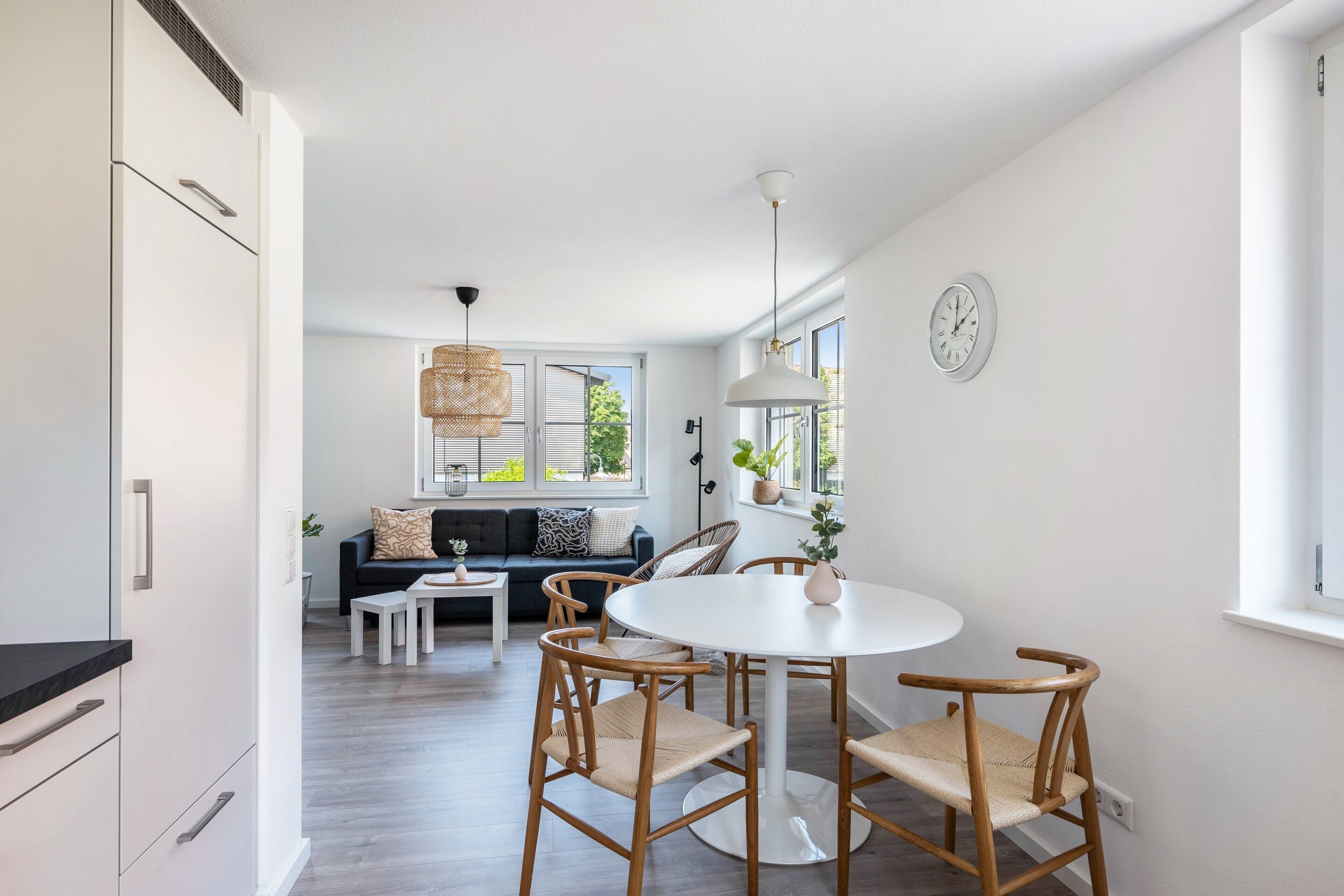 Une cuisine moderne avec des meubles blancs, un coin repas avec une table ronde et des chaises en bois, et un salon avec un canapé et une grande fenêtre.
