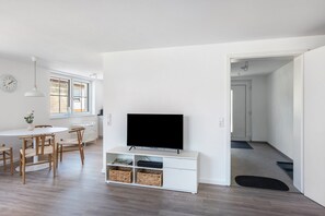 A modern living room with a flat-screen TV, a dining area with a white table and chairs, and a kitchen in the background. - Apartment at the Fountain (Schrozberg)