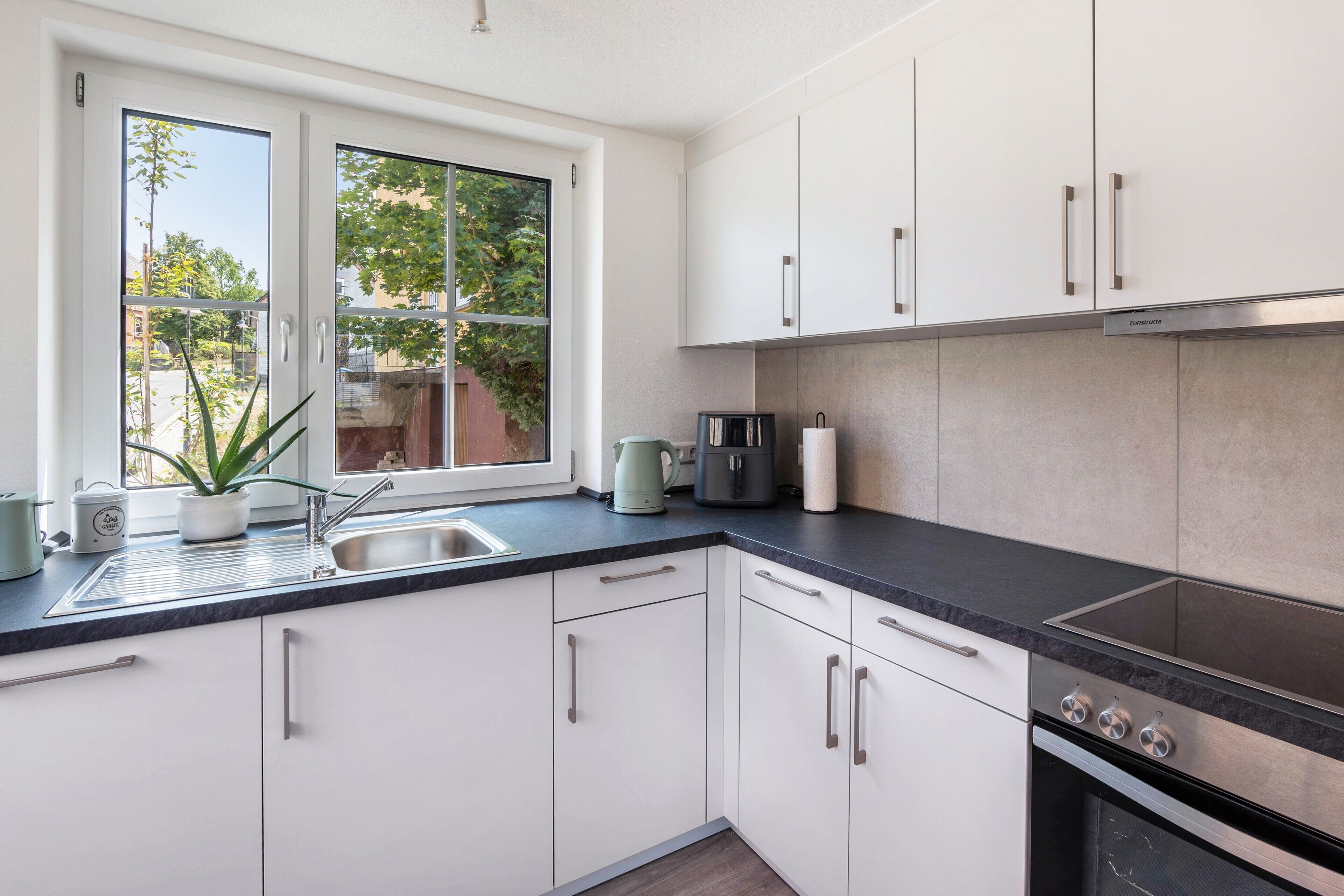 Eine moderne Küche mit weißen Schränken, einer schwarzen Arbeitsplatte und einem Fenster mit Blick auf einen Garten.
