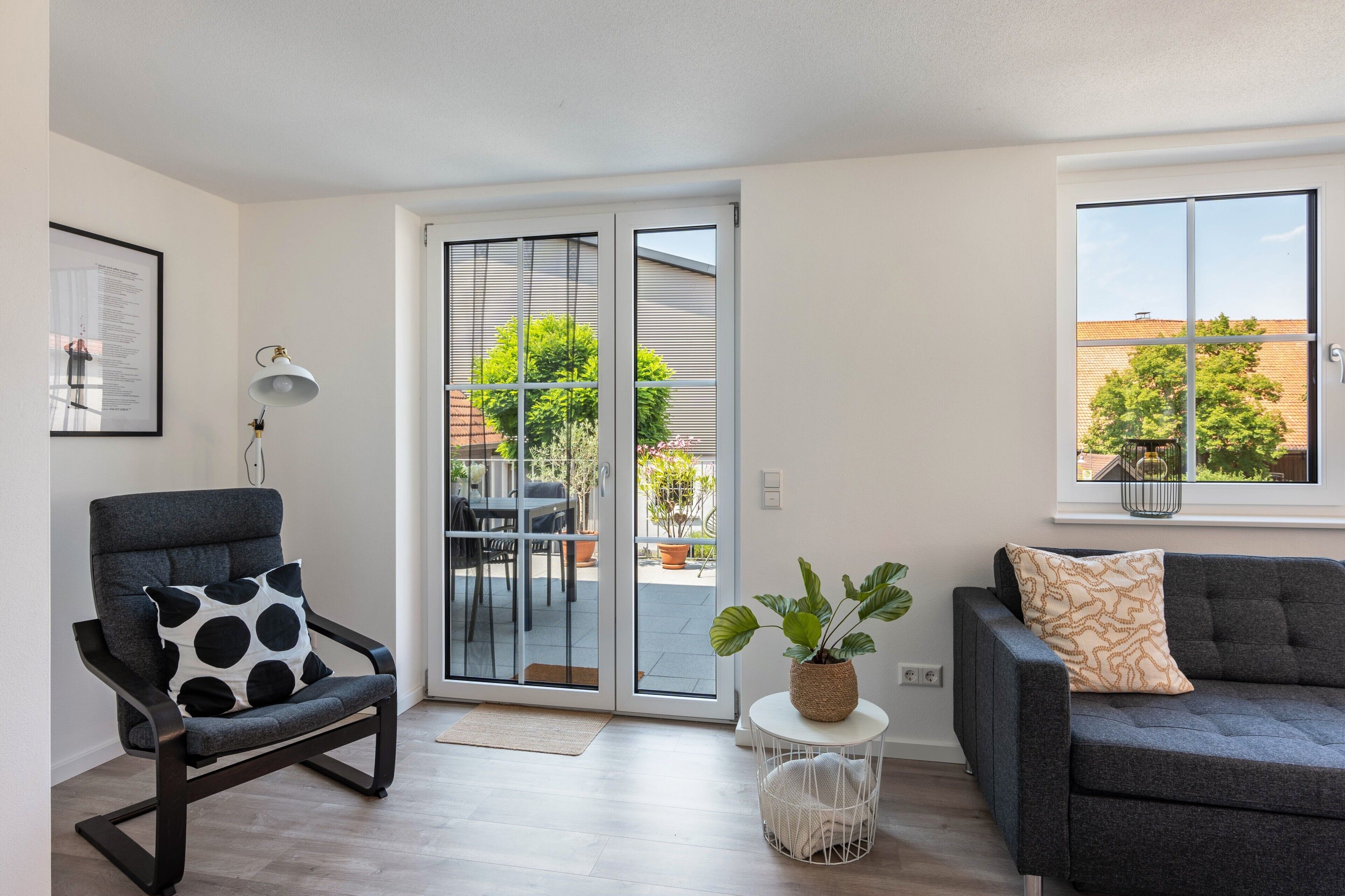 Un salon moderne comprenant un canapé, un fauteuil et une table à manger avec des chaises. Une porte-fenêtre coulissante donne accès à un espace extérieur agrémenté de plantes et offrant une vue sur le quartier.