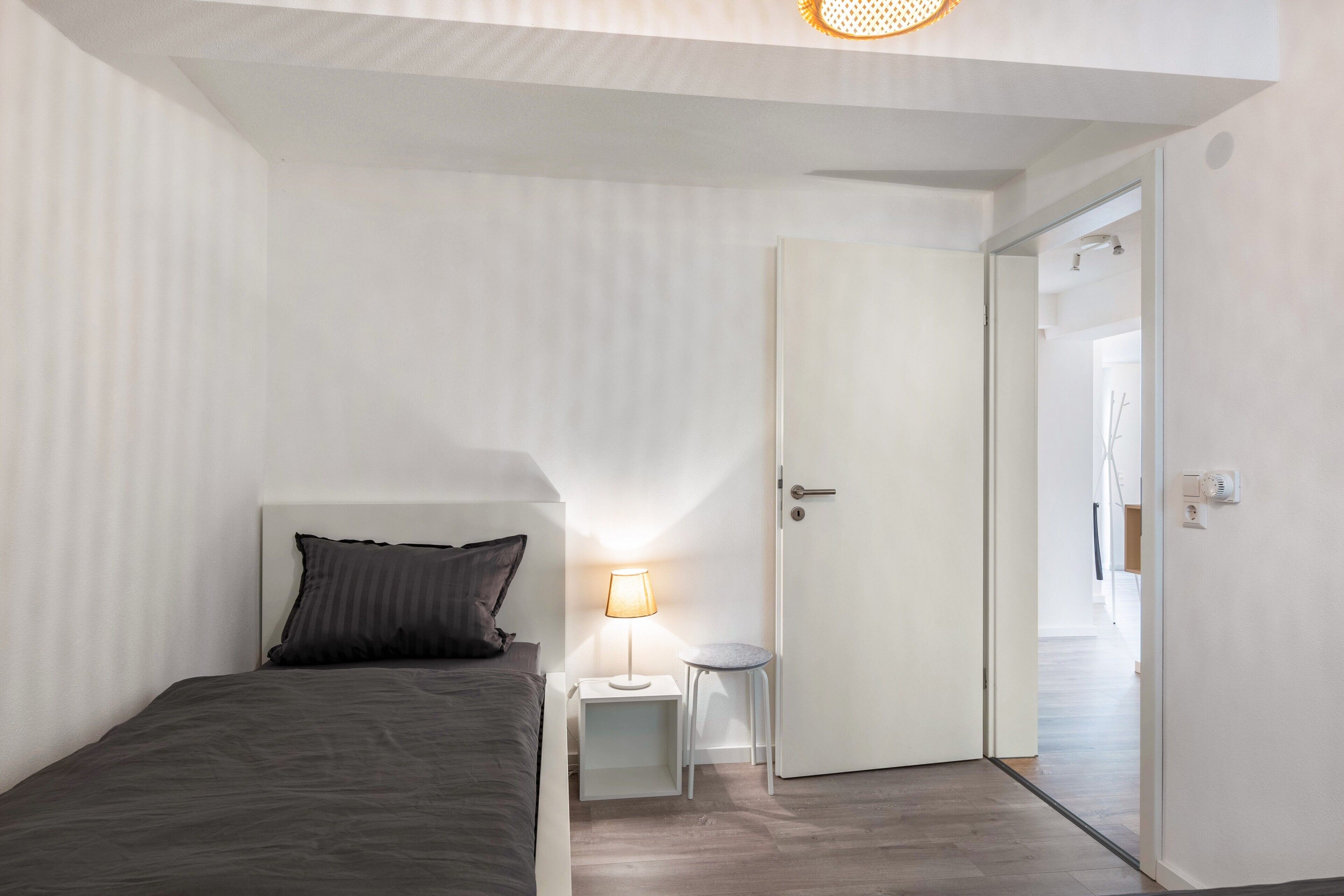 Ein Schlafzimmer mit einem Bett, einem Nachttisch mit Lampe und einer Tür zu einem weiteren Raum.