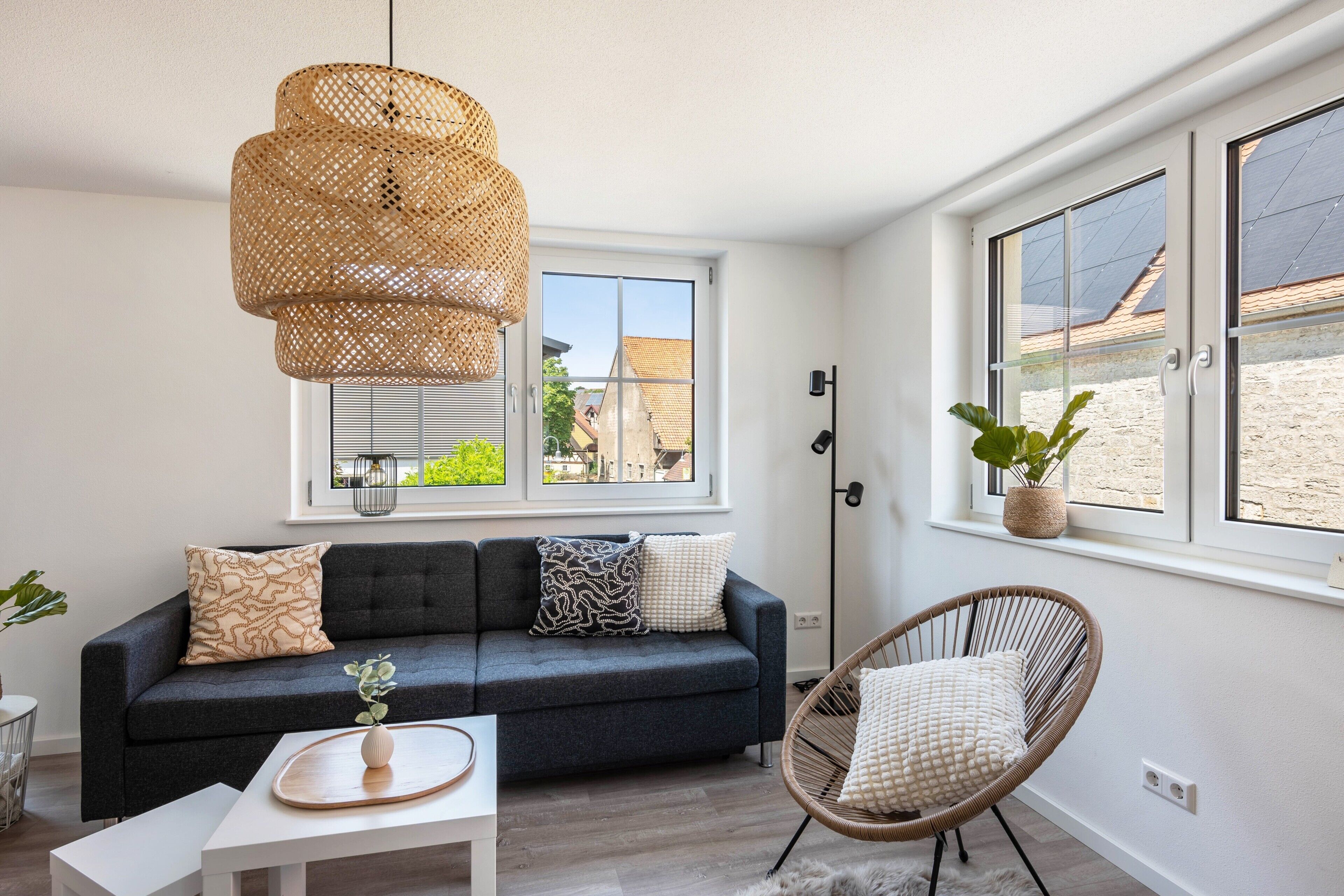 Un salon moderne avec un canapé gris foncé, un fauteuil en osier, une table basse blanche et une grande fenêtre donnant sur des immeubles.
