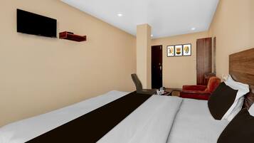 A hotel room with a bed, a TV mounted on the wall, a desk with a chair, and a red armchair.