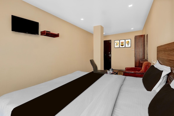 A hotel room with a bed, a TV mounted on the wall, a desk with a chair, and a red armchair.