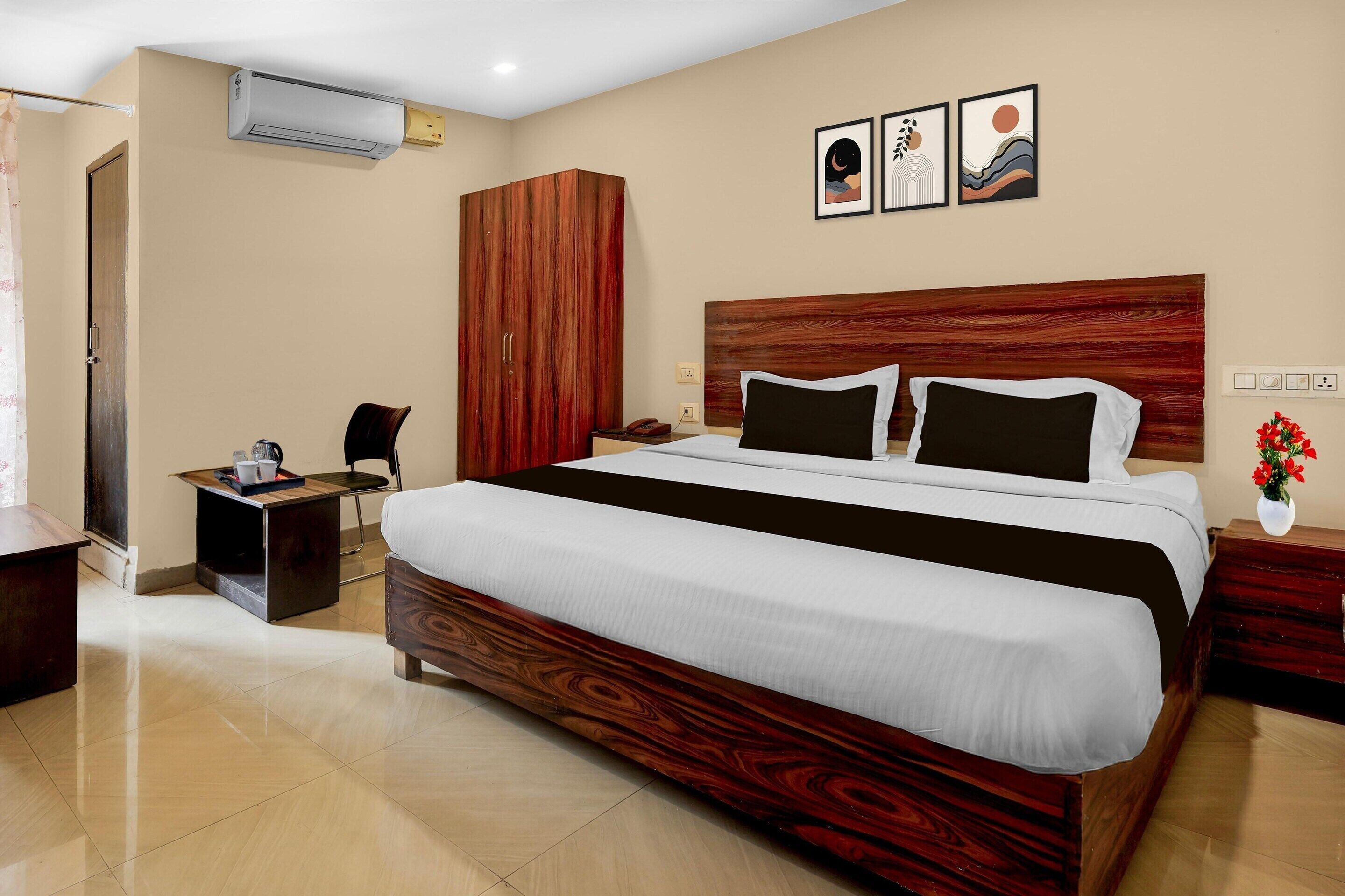 A hotel room with a large bed, a wooden wardrobe, a small desk with a chair, a bedside table with a vase, and a wall-mounted air conditioner.