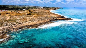 A coastal landscape with a rocky shoreline, clear blue waters, and a distant town. - Luxury 5BR Villa in Paphos | Private Pool, Hot Tub & Sea Views | Family Retreat (Kissonerga)