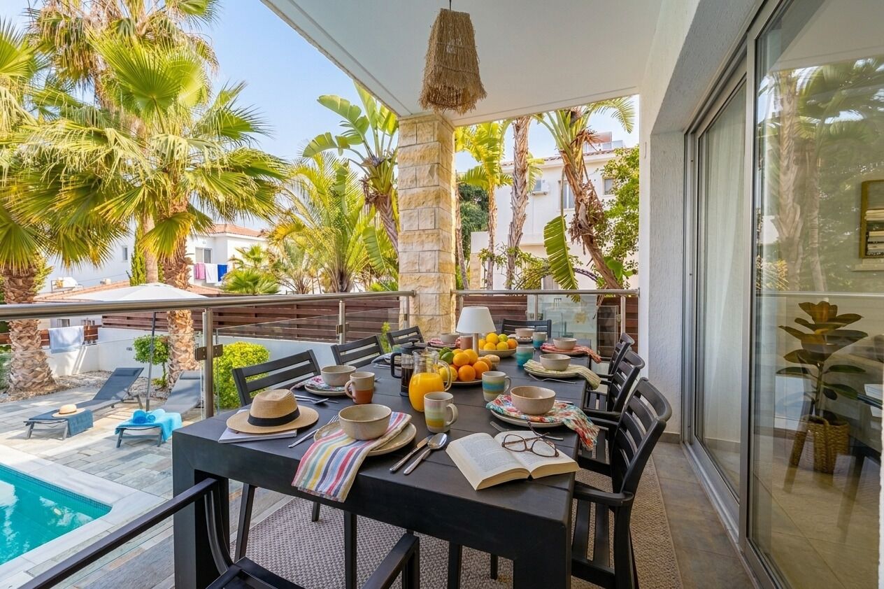 A dining area with a table set for two, surrounded by palm trees and a pool.