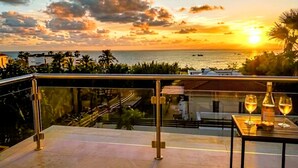 A balcony with a glass railing overlooking a sunset view, a table with two glasses and a bottle, and a distant view of the sea and buildings. - Luxury 5BR Villa in Paphos | Private Pool, Hot Tub & Sea Views | Family Retreat (Kissonerga)