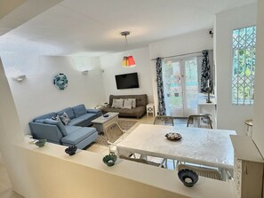 A living room with a sofa, a TV, a dining table, and a balcony with curtains. - Secure & Peaceful Residence – Beach 600m & Nearby Amenities (Flic-en-Flac)