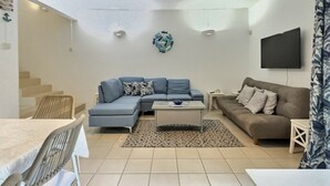A living room with a sectional sofa, a smaller sofa, a coffee table, a TV mounted on the wall, and a staircase leading to another level. - Secure & Peaceful Residence – Beach 600m & Nearby Amenities (Flic-en-Flac)