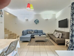 A living room with a sectional sofa, a smaller sofa, a coffee table, a TV mounted on the wall, and a staircase leading to another level. - Secure & Peaceful Residence – Beach 600m & Nearby Amenities (Flic-en-Flac)