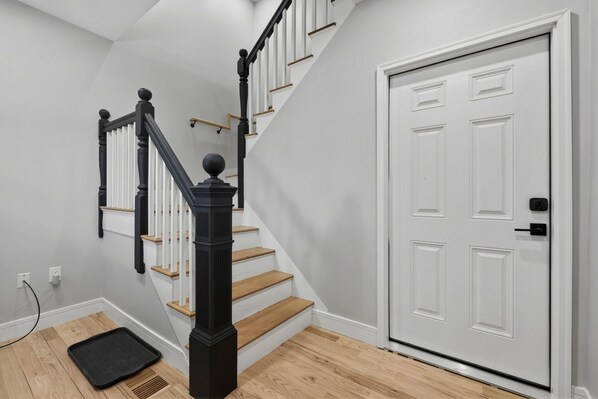 A staircase with wooden steps and black balusters, a white door with a handle, and a black doormat on the floor. - La Oasis: Entire Luxury SFH w 5 BR/3 BA near BOS (Everett)