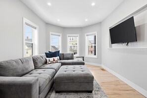 Un salon moderne avec un canapé d’angle, un téléviseur à écran plat fixé au mur et de grandes fenêtres.