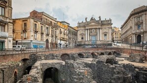 Uma antiga ponte de pedra com arcos, um caminho serpenteando por entre ruínas e edifícios clássicos ao fundo.