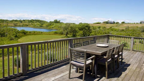 Terraza de madera con mesa y sillas, con vista a un lago y follaje.