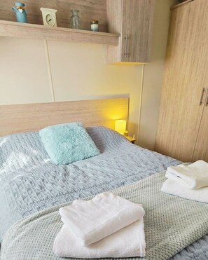 Ein ordentlich bezogenes Bett mit einem flauschigen Kissen, einer Nachttischlampe und einem Stapel gefalteter Handtücher.