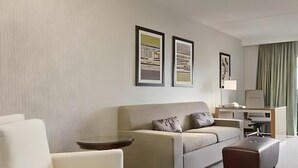 A modern living room with a sofa, armchair, and ottoman, a desk with a lamp, and framed artwork on the wall. - Embassy Suites By Hilton Auburn Hills (Auburn Hills)