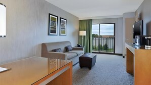 A hotel room with a sofa, a small table, a TV, a balcony with a view, and framed pictures on the wall. - Embassy Suites By Hilton Auburn Hills (Auburn Hills)