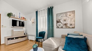 Een moderne woonkamer met een flatscreen televisie, een bank, een fauteuil, een bijzettafeltje met een plant en een groot bloemschilderij.