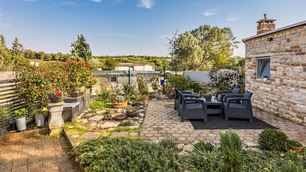 Een stenen gebouw met een bakstenen schoorsteen, een geplaveid terras met tuinmeubilair en een tuin met verschillende planten en een hek.