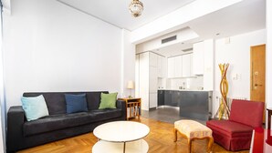 Ein modernes Wohnzimmer mit einem schwarzen Sofa, einem runden Couchtisch und einem roten Sessel. Im Hintergrund ist eine Küche mit weißen Schränken und einer Marmorarbeitsplatte zu sehen.