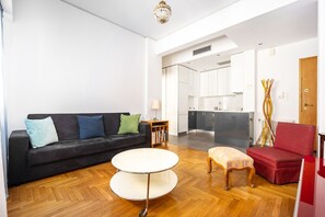 Sala de estar moderna com sofá preto, mesa de centro redonda e poltrona vermelha. Uma cozinha visível ao fundo apresenta armários brancos e bancada de mármore.