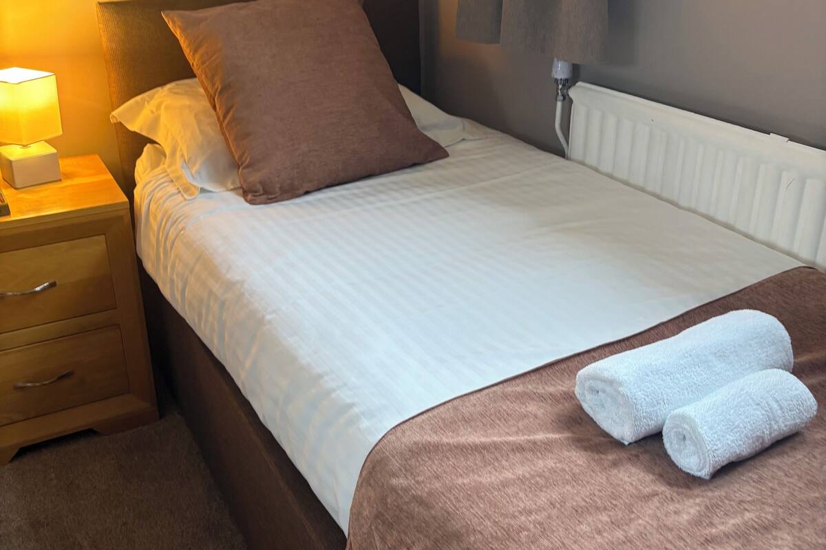 A neatly made bed with a brown headboard, a bedside lamp, and a pair of rolled towels on the bed.